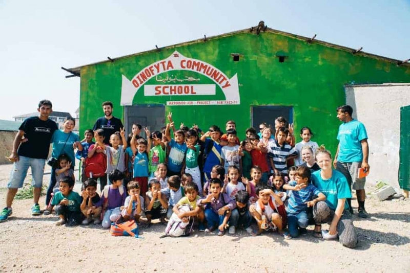 Voluntaris de l'ONG Armando Aid amb els infants del camp d'Oinofyta. Al darrera, l'escola. Autora: Pilar Liberty Munro.